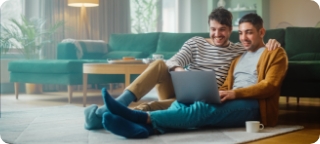 Couple sits on floor of apartment, looking at a laptop together.